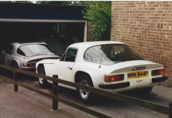 M Series Fuel Tank - Page 1 - Classics - PistonHeads - The image shows a tranquil scene featuring two vintage cars parked side by side in a garage. The cars appear to be a vintage grey model and a white model with the license plate readable as "MHN 84P". They are parked in a spot bordered by a wooden fence with a wire running along the top. The setting suggests a residential garage area with a brick wall visible in the background. The overall mood of the image is serene, with the vintage cars as the focal point amidst the quiet surroundings.