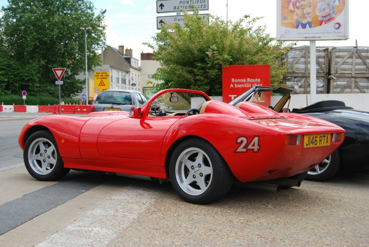 G26 better than G33 ... ? - Page 1 - Ginetta - PistonHeads - The image shows a vibrant red convertible car parked on the road side. The car is positioned diagonally, facing the left side of the image. A person is visible driving the car, adding a dynamic element to the otherwise static scene. In the background, there are two other cars parked appropriately. The road itself appears to be in a bustling area, with street signs of various colors and shapes, adding depth to the scene. The overall atmosphere of the image is lively and suggests that the photo was taken during the day.