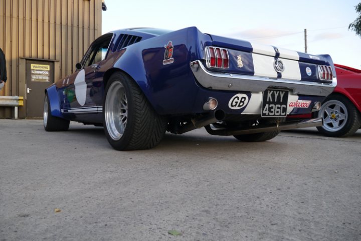 The GT Cafe - the ace cafe of the north - Aug meet - Page 1 - North West - PistonHeads - The image shows a vintage muscle car parked on a paved surface. It is a brightly colored two-door vehicle with a custom paint job, featuring a combination of dark navy blue and silver. The car has a distinctive hood, commonly associated with classic muscle cars. The license plate displays a combination of letters and numbers, and there are at least two people in the background, one of whom appears to be standing near a bench. The setting suggests a casual, outdoor environment, possibly a driveway or parking lot.