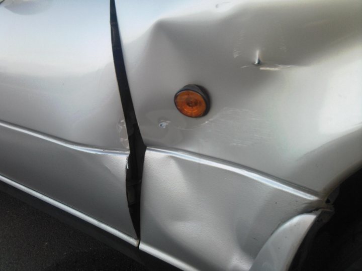 Pistonheads Booked Bodyshop - The image shows a rear view of a silver car with a significant dent on the side near the back. The silver bodywork is visibly damaged, with a noticeable crease and ripples around the dent. The car also appears to have a small round emblem on the rear, which is typical for taillights or bumpers, but it is obscured by the damage. The area around the dent appears to have bald patches, suggesting that it may have been recently repaired or is in need of repair. The image could be used for artistic or real-world contexts that require visual representation of a damaged car.