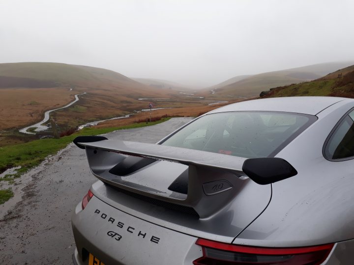 Flipped GT3.2's - Page 28 - 911/Carrera GT - PistonHeads - The image features a Porsche parked on the side of a road, overlooking a lush green valley under an overcast sky. The car is positioned facing the camera at a slight angle, showcasing the rear with its spoiler partially visible. The passenger seat window is slightly down, reflecting some of the cloudy surroundings. A winding road with a drainage system on one side leads the eye into the lower left part of the image.