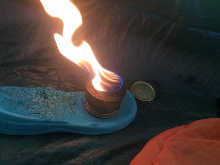 Tent,mountains, freezing, trangia - Page 1 - Tents, Caravans & Motorhomes - PistonHeads - The image showcases a candle with a flame at the top, positioned on what appears to be a blue object, possibly a candle holder. This object is placed on a textured, grey surface, which could be a fabric or a mat. Additionally, there are two coins seen in the image, one of which is located directly behind the candle, while the other coin is visible on the right side of the image. The coins and the candle seem to be the primary subjects in this image.