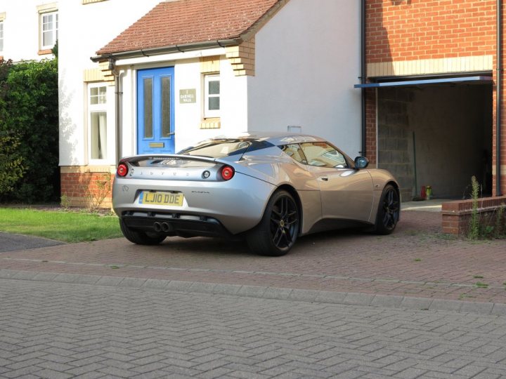 I've also bought one.... - Page 1 - Evora - PistonHeads - The image shows a silver sports car parked at the side of a narrow residential street. The car's sleek design and shiny detailing suggest it is a newer model. In the background, there are a couple of two-story houses with white walls, one with a blue door, and a garden with trees and bushes. The driveway where the car is parked seems to be made of brick pavers. The overall scene is quiet and residential.