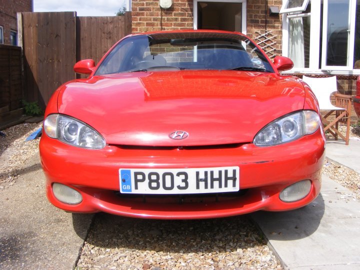 Coupeshed Pistonheads - This image shows a vibrant red Hyundai car parked in the right side of the frame, slightly angled to the viewer. The car's license plate is prominently displayed. The setting appears to be a residential area, as there are fences and a ladder visible in the background, suggesting a backyard or similar private space. The weather seems mild, as there are no indications of extreme temperatures or conditions. The car itself is in good condition, with a clean exterior and well-maintained tires.