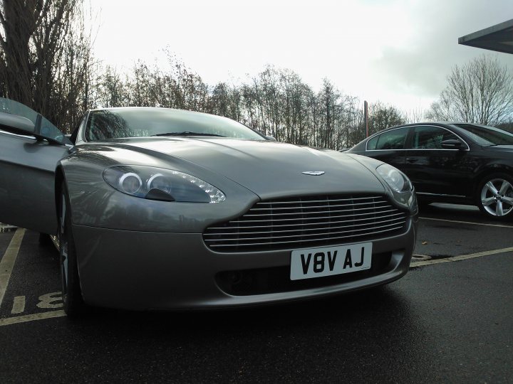 Anyone use their Aston? - Page 2 - Aston Martin - PistonHeads - This is an image of a luxurious, silver-colored car parked on a wet street. The car is an Aston Martin, elegantly positioned in a parking lot with another vehicle, possibly a black Audi car, parked behind it. The Aston Martin is near a black car, indicating they may be parked close together. The sky in the background is overcast, suggesting that it might be raining or have rained recently. The tires of the car are also visible, reflecting the wet surface of the street.