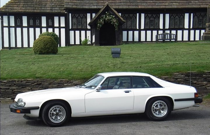 What "digitally remastered" classic would you like to own? - Page 5 - General Gassing - PistonHeads - The image shows a white classic car parked in a driveway in front of a traditional British house. The car appears to be an older model, possibly a British sports car, its sleek white body polished and gleaming under the light. The house is a charming Tudor-style structure with white-painted walls, pointed roof, and a front garden ending in a green lawn. There are potted plants and garden decorations visible in the front yard. The car is in the shade, suggesting it's possibly daytime with some cloud cover. There are no texts or branding in the image.