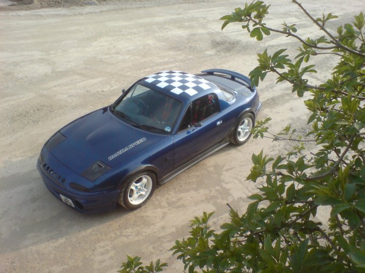 Pistonheads Show - The image shows a sleek, dark blue sports car with a black and white checkered pattern on its roof, parked on a gravel lot. The vehicle is a two-door convertible, with the top slightly down, allowing a partial view of the passenger seat. The surrounding area is mostly bare dirt, and there are green leaves in the foreground of the image. There is no visible text or branding in the photo. The style of the image is a naturalistic photograph with a focus on the car.