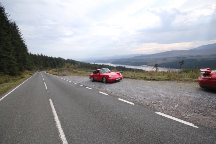 Highlands - Page 69 - Roads - PistonHeads - In the image, two red Porsches are parked at the side of a winding, paved mountain road. The car on the left is positioned slightly closer to the road, while the one on the right is further into the shoulder. Both cars are facing the camera, pointing towards a distant view of a river flowing through a valley, with mountains in the background, visible under a partly cloudy sky. The road itself is marked with white lines on either side, indicative of road markings. The overall setting suggests a scenic drive in a mountainous area.