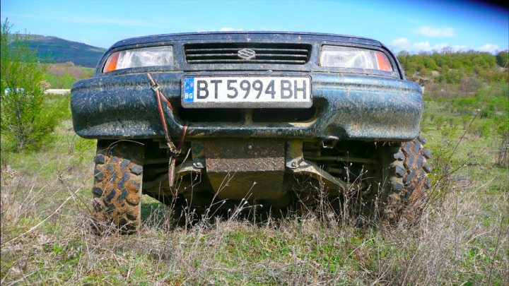 Home made underbody protection - Page 1 - Off Road - PistonHeads - The image captures an abandoned or off-road black sedan with mud all over it. The vehicle is missing its front bumper and hood, revealing portions where the bodywork has been damaged. Over the license plate, the registration number "BT594H" is clearly visible. The car is parked in a lush green field or meadow, surrounded by tall grass and a few scattered plants, and stands against a backdrop of a clear sky and a distant mountain or hill.