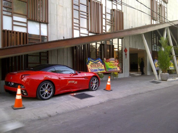 - Page 431 - Supercar General - PistonHeads - The image shows a city street scene with a red sports car parked in front of a building. The sports car has a convertible top, which is partially back, revealing some of the interior. It features chrome details and alloy wheels. There are a few potted plants along the sidewalk, adding to the urban atmosphere. The building has a modern architectural style, and there are awnings extending from the facade. A person is visible in the background, indicating some activity within the building.