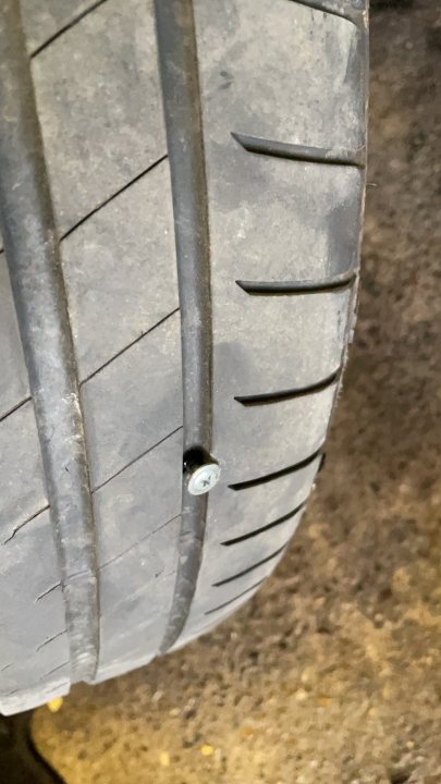 The 'Oh crap, I won it' E39 530i - Page 9 - Readers' Cars - PistonHeads UK - The image shows a close-up of a car tire with a visible bolt. The tire tread is worn, indicating it may be old or frequently used. In the background, there is a glimpse of what appears to be a garage or workshop setting with tools and other equipment that suggests an environment for vehicle maintenance. The focus on the tire's condition implies a concern for safety or performance, possibly in relation to the car's performance or impending service.