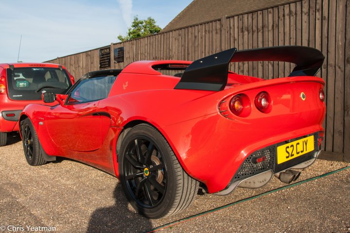 A red motorcycle parked in a parking lot - Pistonheads - The image depicts a vibrant red sports car parked in front of a wooden fence. The car's body gleams with a meticulous shine, accentuating its aerodynamic design and wide stance. The air vents on the car's rear are prominent, contributing to its sporty appearance. In the background, a red SUV is partially visible, providing a contrast in size and purpose to the sports car. The setting appears to be a parking area with a smooth road leading to the fence, under a clear sky.