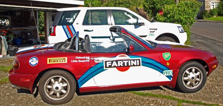 Stickering up for 2016 - Page 1 - Le Mans - PistonHeads - In the image, a red sports car is prominently parked on a gravel driveway. The car is highly decorated with various stickers and flags on its hood and body. The setting is residential, with a white house and a parked white truck in the background. The driveway is bordered by grass on one side and a fence on the other, providing a sense of seclusion. The red car's vibrant color and decorative elements draw the viewer's attention immediately upon viewing the image.