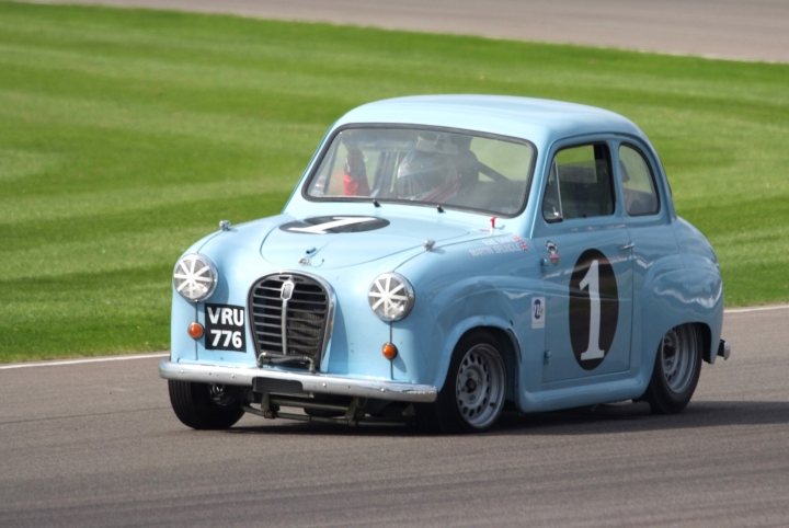 Brundle Pistonheads Austin - The image showcases a scene on a racing track. At the heart of the scene is a vintage blue racing car, bearing the number "1", that's in motion on the track. The car, boasting the number "7" and other racing insignia, displays a classic design. The racing car is positioned slightly off the center of the track, leaning into a right turn as it continues its race. Surrounding the track, a vibrant green landscape stretches out, providing a picturesque backdrop to this high-performance scene. The image captures a moment of intense competition in a classic racing setting.