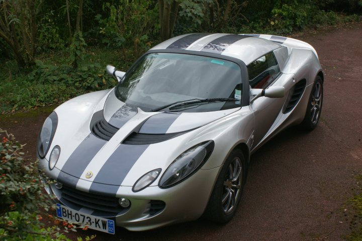 Pistonheads - The image features a silver sports car parked on a dirt road. The car is adorned with a striking black and white stripe running down its center, adding a sporty touch to its design. The car, parked on the left side of the road, is facing towards the right side of the frame. The driver's side is visible in the frame, with the side mirror and window allowing a clear view into the cabin. The surrounding environment is lush with greenery and vegetation, creating a tranquil and picturesque setting for the vehicle.