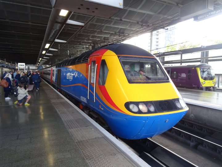 Pistonheads - The image depicts a vibrant scene at a train station. A red and white train, labeled "First," is prominently positioned on the right side of the frame. People are scattered around the platform, some standing close to the edge, while others are further away, giving a sense of depth and activity. To the left of the image, another train can be seen, hinting at multiple routes or directions. The station itself is well-lit, with natural light streaming in from the windows above. There's no text present in the image. The overall scene conveys a typical day of commuter traffic at a train station.