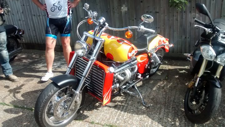 The "Photos From Today's Ride" thread... - Page 168 - Pedal Powered - PistonHeads - The image features an exceptionally colorful motorcycle, predominantly in shades of orange and red, parked on a textured surface. The motorcycle is detailed with numerous orange, silver, and black parts, as well as a yellow body. One light opens up the front, revealing a headlight and a starimg engine underneath. There is a sense of a vibrant garage environment filled with the passion for motorcycling. The backrests of two other motorcycles can be seen, which appear darker in color.