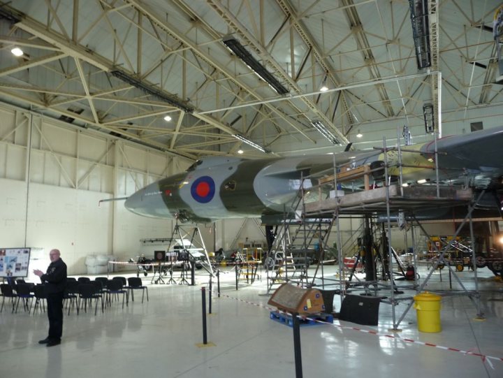 Pistonheads - The image features a large airplane suspended from a high ceiling inside a hangar. Its undercarriage is more visible on the left side as the plane stands on a steel frame. The hangar has a sturdy structure with metal support beams crisscrossing the ceiling. There's ample open space around the plane, and various equipment and parts are scattered about, suggesting ongoing maintenance or preparation for flight. A view of the airplane's top can be seen from the side, allowing one to identify the British roundel markings.