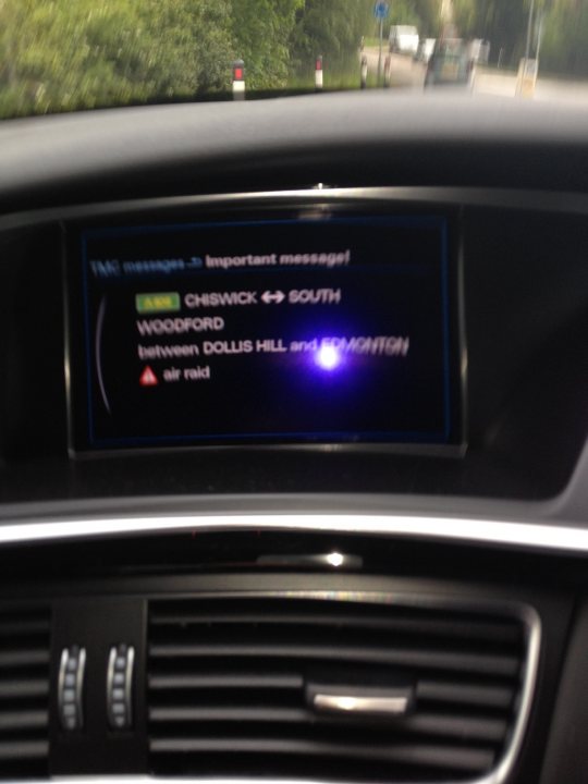 A close up of a tv with a digital screen - Pistonheads - The image appears to capture a car's computer screen as seen from the passenger seat, showcasing important message-related notifications. The top portion of the screen displays an electronic message indicating the vehicle has reached "Chiswick or South" and is nearing an intersection. Below that, there is a message regarding "Woodford." A green light is displayed above the bottom portion of the screen. On the bottom right side of the screen, near the top of the image, there is a red triangle signaling an air-raid alarm. The car's interior lights are on, hinting at the time of day.