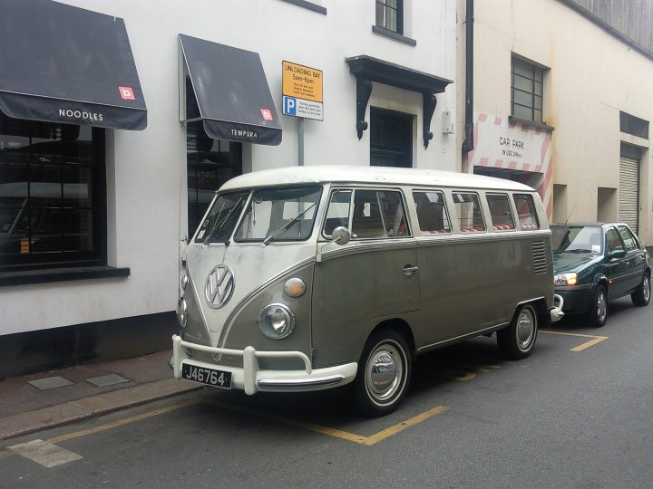 COOL CLASSIC CAR SPOTTERS POST!!! Vol 2 - Page 64 - Classic Cars and Yesterday's Heroes - PistonHeads - In the image, a vintage Volkswagen bus is the main focus. The bus is white and parked next to the sidewalk on the side of the street. It has a large window on the back and a license plate that reads "J6 R764". The bus is near a building with white walls, a metal awning, and a blue sign with Asian characters on it. Additionally, there's a grey car parked opposite the bus, adding to the small-town street scene.