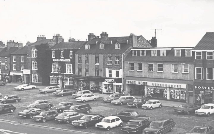 A 'period' classics pictures thread (Mk III) - Page 43 - Classic Cars and Yesterday's Heroes - PistonHeads UK - The image is a black and white photograph that captures an urban setting. It features a parking lot filled with various vehicles, including cars and trucks, suggesting a bustling area. The scene includes buildings in the background, which may be commercial or residential structures. A row of parked cars lines the foreground, indicating a street adjacent to the parking lot. The absence of color in the image gives it a timeless quality, emphasizing the architectural and structural elements within the composition.