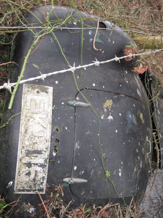 Any ideas what it is? - Page 1 - Classic Cars and Yesterday's Heroes - PistonHeads - The image features a large black stump in a field, close to the camera. Barbed wire stretches across the edge of the stump. A license plate, showing only the letter "K", is partially visible on the stump. The stump is covered with a quantity of dirt and the barbed wire atop it appears to be from a former fence. The scene conveys an atmosphere of abandonment or neglect.