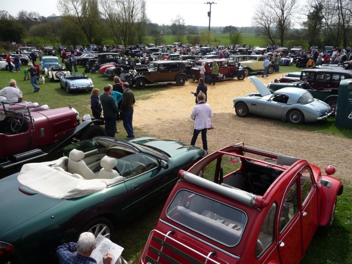 Pistonheads Showtime Boys Bentley - The image shows a large open field with a vast array of various models and colors of vintage cars and trucks parked in rows and along the periphery. The vehicles, representing different eras, vary from small to large with distinctive designs such as clear windshields, convertible tops, and classic tail fin styles. The sun is shining, and the grass is green, suggesting a warm day. A number of people are scattered throughout the scene, inspecting the vehicles, walking among them, and engaging in discussion groups around the cars. The scene appears to be a car show or a gathering of vintage vehicle enthusiasts.