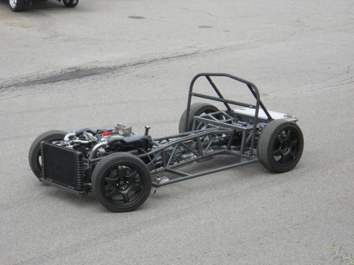 5000M build - Page 1 - Classics - PistonHeads - The image shows a homemade, lightweight race car parked on an asphalt surface. The car has a custom roll cage and features two small black wheels on its front and two larger wheels at the back. It's equipped with a flat black panel at the rear for weight distribution and aerodynamics. The open chassis suggests a kit car, and there's no visible engine or bodywork. The background has other vehicles parked, indicating that this might be a garage or a display area. The car is quite narrow and appears to be ready for a race or track day.