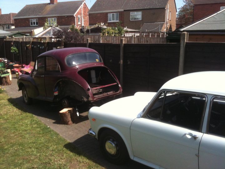 1971 Morris Minor 1000 Resto thread (Maybe) - Page 1 - Classic Cars and Yesterday's Heroes - PistonHeads - The image showcases a suburban scene with two vintage cars parked on the driveway. The car in the foreground is a striking red and black, while the second car, parked behind, is a white Vauxhall. The driveway is lined with a high wooden fence. In the background, houses and a fenced garden enclosed by a metal gate are visible. Considering the lush greenery, it is likely a sunny day, which enhances the appearance of the entrust cars. The image evokes a sense of nostalgia for the golden age of automobiles.