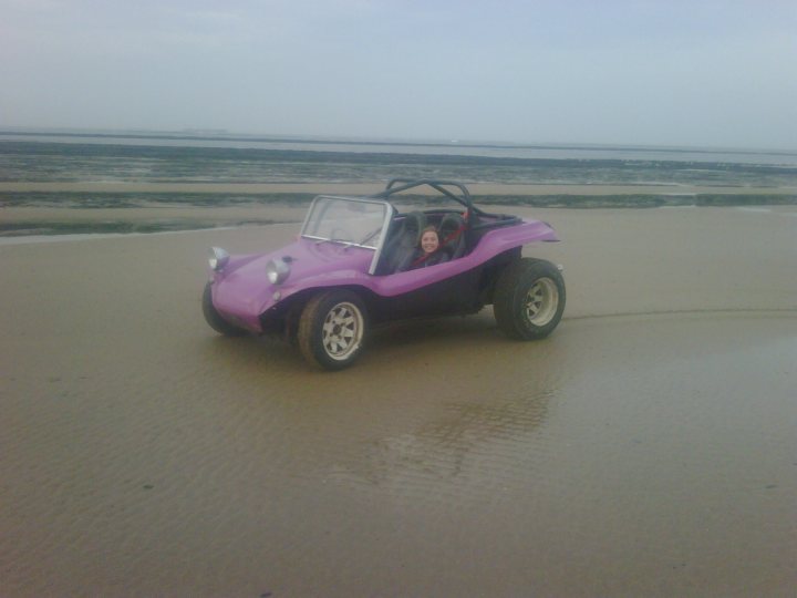 Shine Pistonheads Sun - The image captures a playful scene on a beach with wet, sandy terrain. A girl is sitting in the driver's seat of a custom-made dune buggy, which combines the features of a trikele and a classic car. The buggy is predominantly pink with black wheel wells and white personal plates on the back. The buggy's tires appear to be sturdy and suitable for the sandy conditions. The girl is wearing casual clothing and seems to be driving or possibly going for a spin, adding a sense of adventure to the image. The scene is set against a flat, sandy expanse with a hint of the sea in the distance.