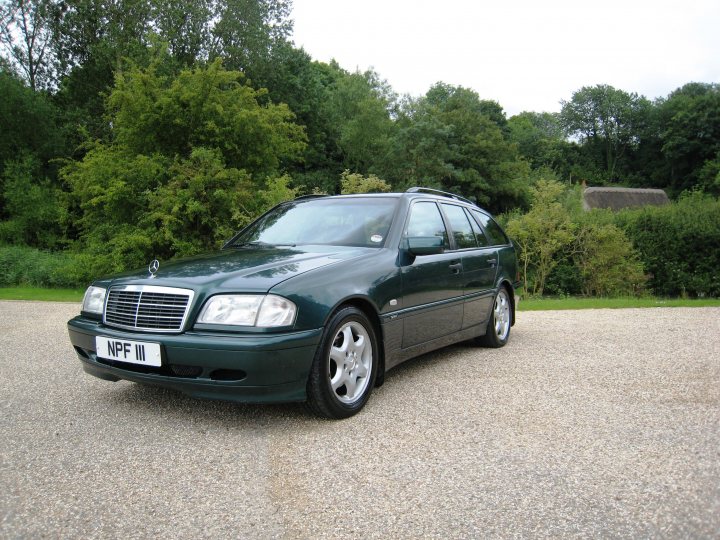 What private plates do you have? - Page 68 - General Gassing - PistonHeads UK - The image shows a green sedan parked on gravel. The car is facing towards the left side of the image. It has a European-style license plate and appears to be in good condition. In the background, there are trees and a house, indicating that the car is parked in a suburban or rural setting. There are no people visible in the scene.