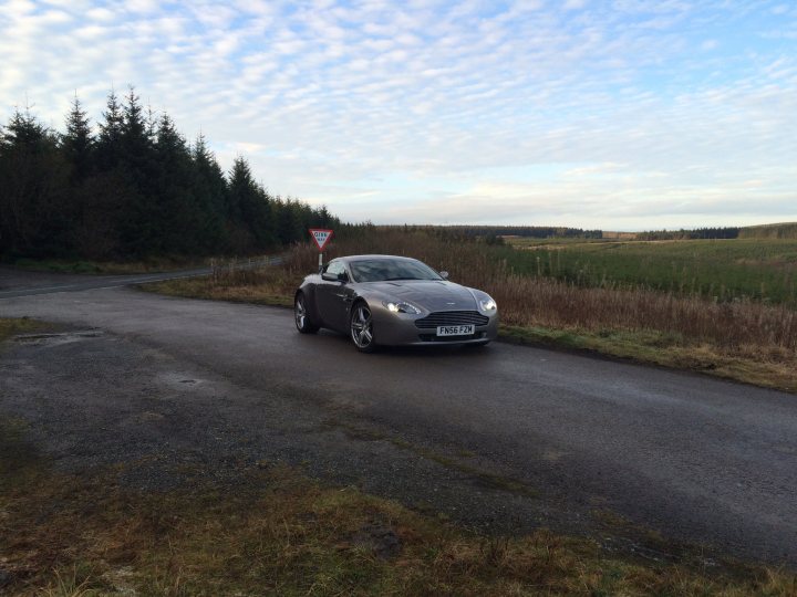 How about an Aston photo thread! - Page 107 - Aston Martin - PistonHeads - In the image, a silver sports car is parked on a winding country road. The car is facing towards the camera, its sleek design contrasting sharply with the natural surroundings. The road itself curves gently to the left, flanked on both sides by grassy fields. In the far distance, several trees can be seen, adding a touch of wilderness to the scene. The sky overhead is a serene blue, dotted with fluffy white clouds, complementing the sense of tranquility and solitude in the scene.