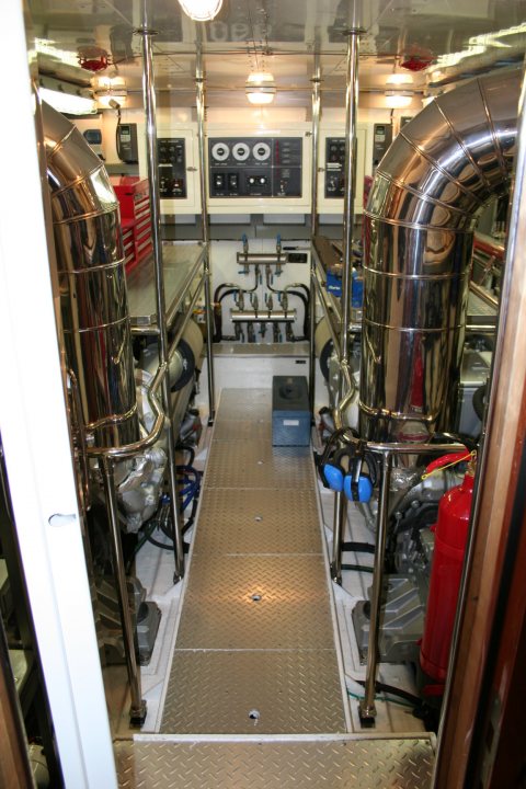 Bleeding Pistonheads Radiator - The image shows the interior of a large industrial or possibly a sports car workshop. The viewer's perspective is as if they are standing on a tiled floor at the opening of a service bay, looking down a corridor to the counter and beyond. The space is filled with a variety of mechanical and industrial equipment, including large steel beams, machinery with metallic surfaces and red fire extinguishers, and numerous wires and tubes connecting different parts. The walls feature black painted control panels with white lettering, and the overall setting suggests a professional environment designed for rigorous use and possibly manufacturing or repairing vehicles given the presence of the fire extinguishers.