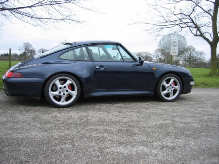 Caution! 911 and CGT content. No residual value chat! - Page 1 - 911/Carrera GT - PistonHeads - The image features a blue classic Porsche parked on a gravel surface. The car is positioned slightly to the right of the frame, with a large clearance behind it. It has distinctive doors that open outwards, a signature of many vintage Porsche models. The cordoned-off background includes a chain fence and what appears to be a satellite dish or water tower. There is no visible text or discernible activities taking place in the image. The style of the image is a real-life photograph with no evident filters or digital alterations.