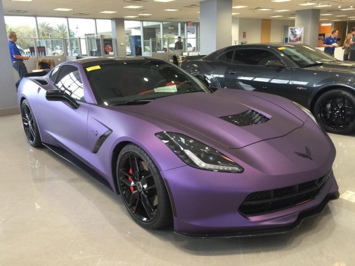 A blue car parked next to a red and white car - Pistonheads - The image showcases a gleaming dark purple sports car parked indoors. It's positioned facing the camera, capturing its front. The car features black rims and round headlights. There are a few other cars in the dealership, but the purple sports car is the main focus. The floor has large windows, letting in ample natural light that highlights the car's unique color.