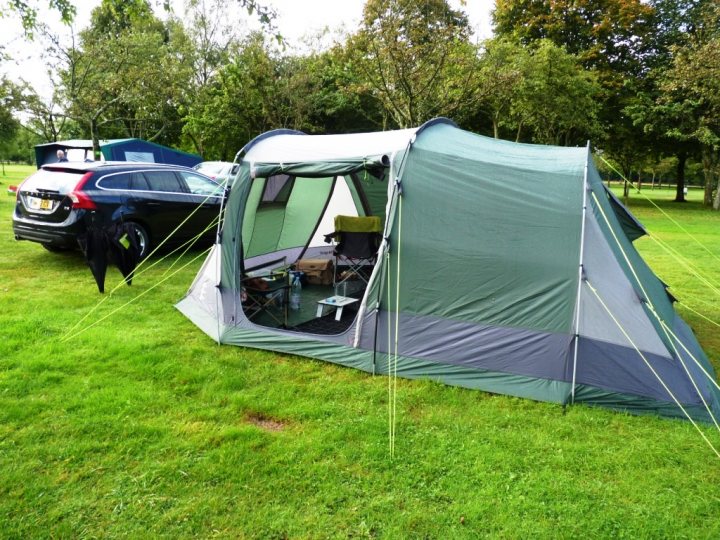 Tent needed - don't know where to start!! Recommendations? - Page 1 - Tents, Caravans & Motorhomes - PistonHeads - The image depicts a green and gray tent set up on a grassy field. A person is seated in a chair inside the open tent, facing the viewer. The tent is partially surrounded by vehicles, including a car and a truck. The backdrop shows a line of trees, and the sky is overcast. The setup suggests a temporary outdoor accommodations such as camping.