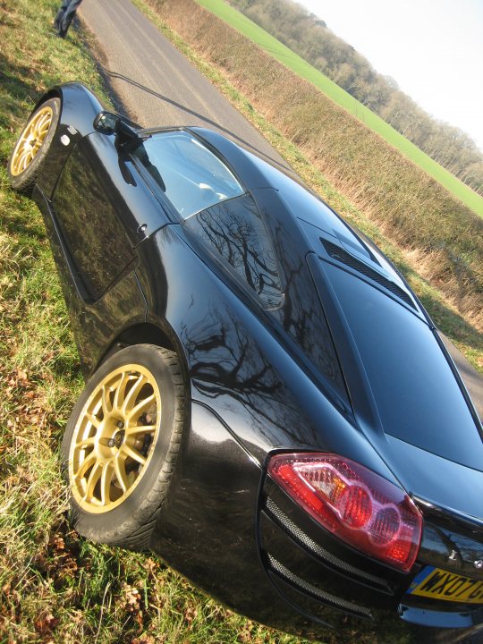 Lotus Europa Gold Wheels - Page 1 - Elise/Exige/Europa/340R - PistonHeads - The image showcases a fast, sleek black sports car positioned on the side of a rural road. The car's design features a prominent ride height differential, with raised wheels on the front compared to the rear, creating a high ground clearance over the rear apron. This differential in height is often seen in high-performance vehicles designed for precision driving and is accentuated by the gold-colored metallic alloy wheels that give a striking contrast to the car's black exterior. The suspicion or idea of a "shiny, beautiful, black car" in the reflection of the lagoon seems to be an incorrect description of this vehicle. The background of the image features a serene, grassy area with trees, suggesting a peaceful, country setting.