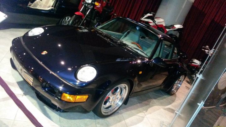 Royal Jordanian Automobile Museum - Page 1 - Middle East - PistonHeads - The image is a photograph featuring a vintage Porsche sports car with a black exterior and yellow headlights, parked indoors. The car is positioned facing the camera, and it appears to be clean and well-maintained. Behind the Porsche, there is another classic motorcycle parked at the rear, partially visible by the motorcycle. The room has a polished floor and a red or maroon curtain to the right side of the vehicle, which provides a complementary background color. The car's model is not visible, but it is a convertible as indicated by the visible soft top roof area. The style of the image is straightforward, capturing a moment with attention to detail and perspective.