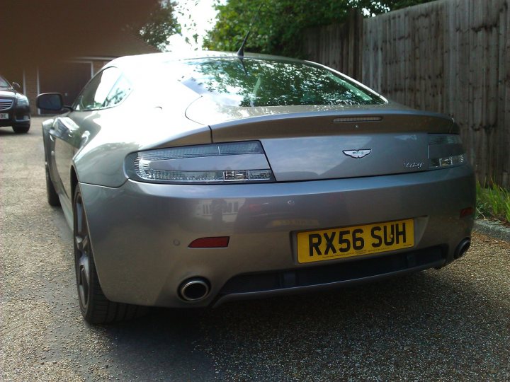 Clear Rear Lights Install - Page 1 - Aston Martin - PistonHeads - This image captures a tranquil scene on a driveway, featuring a striking black Aston Martin DB7 car. The car is positioned at an angle, facing to the right, and is parked on a gravel driveway. The rear end of the car is prominently visible, with a license plate that reads 'RX56 SUEH'. A single tree branches over the scene, adding a touch of nature to the image. The overall color scheme of the image is soothing, with the black car set against a green backdrop. The sunlight shines at an angle, casting gentle shadows and highlighting the car's sleek design.