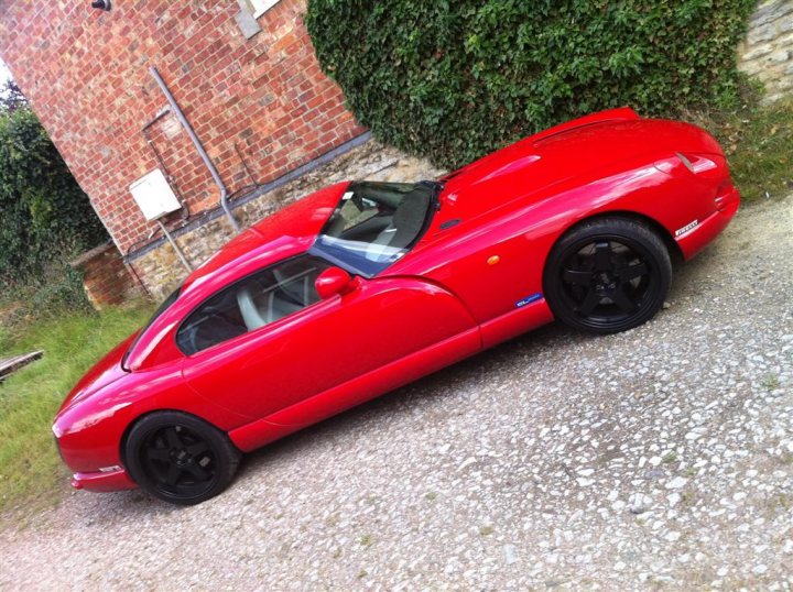 A red car is parked in a parking lot - Pistonheads - The image showcases a vibrant red sports car parked amidst a serene setting. The car is positioned diagonally to the viewer, highlighting its sleek design. A green bush and a brick wall serve as picturesque backdrops to the car, contributing to a tranquil outdoor scene. The vehicle appears to be a convertible model, adding to the overall aesthetic appeal of the car. The red hue of the car stands out starkly against the more muted colors of the backdrop.