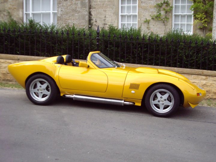 A red car is parked in a parking lot - Pistonheads - The image depicts a striking, yellow Ferrari roadster parked near a stone building with a fence. The roadster has an open cockpit, and its design features a long, sloping hood and rounded, bulging fenders. The tires appear to be made of exposed metal braiding, enhancing the classic sports car aesthetic. The vehicle is parked on a concrete surface with a reflective sheen, implying a shiny, polished surface. The sky is clear with few clouds, suggesting a sunny day.