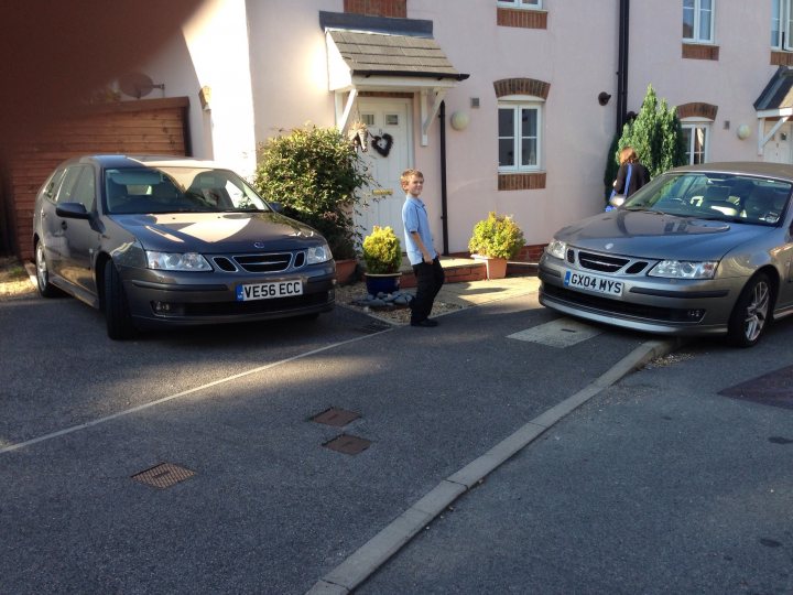 Welcome to Flight School! - Page 1 - Saab - PistonHeads - The image shows two parallel parked cars on a driveway with an open garage door beside them. A young boy, wearing a blue short-sleeve shirt, stands between the cars, appearing to be watching them. The car closest to the viewer has a license plate that reads "V656" and is a darker color. The car parked farther away has a license plate that reads "EX44 NWY." The building behind the cars has several windows with some potted plants visible.