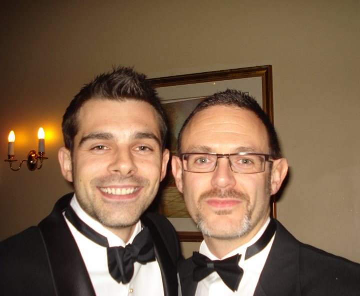 Btap Polaroids Pistonheads - This image is a portrait of two men posing together, likely at a formal event. They are both dressed in formal attire, with each wearing a black bow tie, and one of them sports a mustache. Their poses and expressions suggest a friendly and relaxed atmosphere. In the background, there is a wall-mounted light fixture and a framed artwork hanging on the wall. The men are smiling and seem to be enjoying the moment.