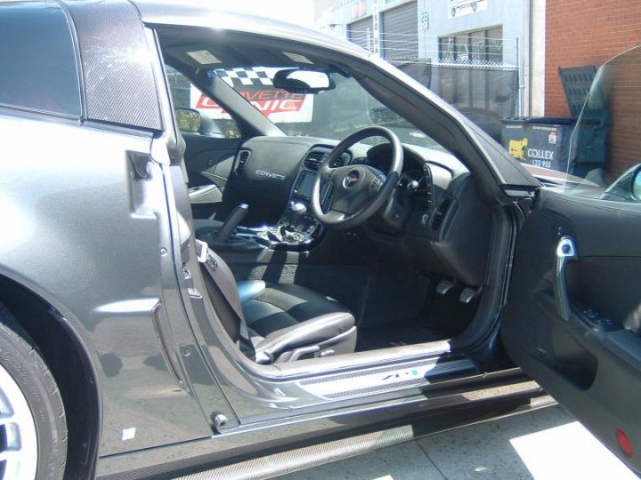 Right hand drive ZR-1 Anybody? - Page 1 - Corvettes - PistonHeads - This is an image of a car, captured from an interior perspective, focusing on the driver's side door. The door is left open, revealing an empty passenger seat. The interior door handles and window regulator are visible, suggesting a tidy and well-maintained vehicle. In the background, the tail end of another car can be seen, suggesting that the photo was taken in a parking lot or similar setting. The clearance of the open driver's door indicates that the vehicle is stationary when the photo was taken.