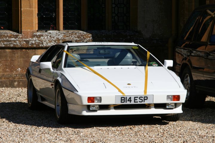 Ideas about value? - Page 1 - Esprit - PistonHeads - The image features a vintage white sports car parked on a gravel driveway. The car is adorned with a yellow ribbon tied and decorated with a green bow on the front. There is another vehicle visible in the background, partially visible inside a garage-like structure that suggests a large, possibly historic building. The setting gives an impression of a formal gathering or an important event.