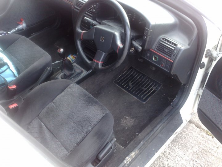 Pistonheads Daily Workhorse Runner - The image captures the interior of a vehicle, particularly focusing on the driver's side. The interior is predominantly black, featuring seats and a steering wheel with red accents. The operation buttons and controls, along with the radio console, are visible behind the steering wheel. The floor of the vehicle is covered with a black carpet, and various items such as a book, bottle cap, and set of keys are scattered around the dash. The perspective of the image suggests it was taken from outside the car, likely from the passenger door area.
