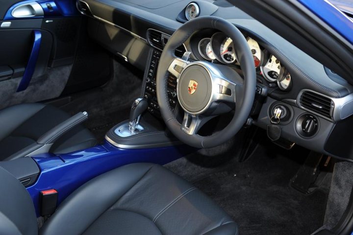 This image features the interior of a luxury car, specifically focusing on the driver's side. A control panel with several dials and buttons is in the foreground. The upholstery of the front seats matches the exterior color, creating a cohesive aesthetic. The car's thorough conditioning shines through its polished surfaces. The steering wheel, a prominent part of the scene, is equipped with a logo, indicative of the brand's signature.