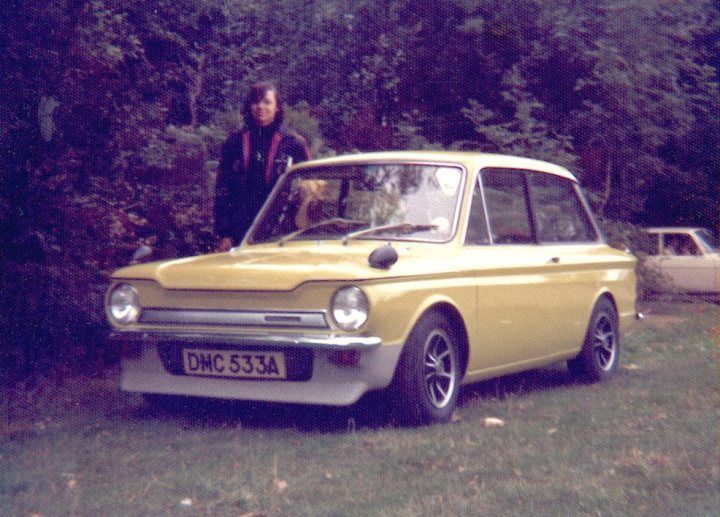 Pistonheads Imp Hillmansunbeamsinger - The image is a photograph featuring a vintage yellow car parked on grass. There's a person positioned next to the car, likely a pedestrian. The vehicle appears to be an older model, perhaps from the 1960s or 1970s, as indicated by its design, such as the round headlights and slim front grille. The front licence plate is clearly visible and displays the letters "DMC". The setting suggests a calm, rural environment, possibly a grassy area alongside a road. The overall mood of the photo is nostalgic, evoking a sense of past travel.