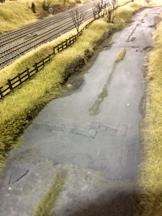 'O' Gauge Model of Heaton Lodge Junction - Page 3 - Scale Models - PistonHeads - The image showcases a scale model of a road and rail intersection. The road appears wet, possibly from rain, with puddles scattered around. The railroad tracks run parallel to the road, both crossing a bridge over a grassy area. The bridge provides an elevated perspective for the image. Solitary trees can be seen near the tracks, adding to the realism of the scene. The overall scene is vintage in style, evoking a sense of nostalgia.