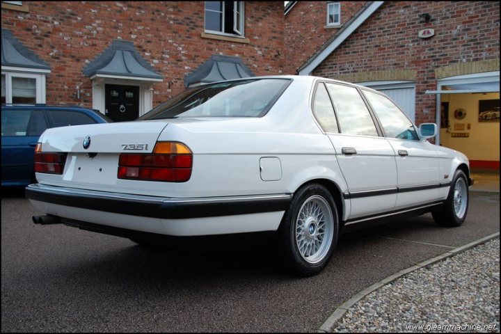 Show Me Your BMW!!!!!!!!! - Page 244 - BMW General - PistonHeads - The image shows a large white BMW parked in front of a brick building. The car's license plate reads "7351." Its tail lights exhibit a classic red color, and it appears to be in good condition. The BMW is situated on a gravel driveway, and there are other cars parked in the background. The building itself exhibits a warm, inviting facade with a partially visible doorframe. The sky is overcast, suggesting the photo might have been taken on a cloudy day.